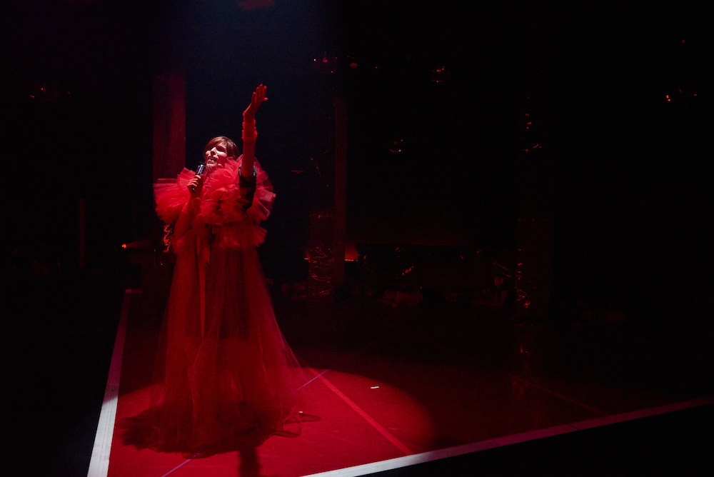 a lone dancer bathed in a red spot light and wearing a slip on gown of fluffy red tulle sings into a hair brush microphone